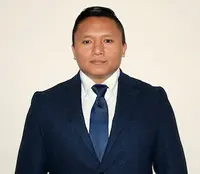 Dhan Gurung stands in front of a light colored wall. He is wearing a dark blue suit and tie, and a white shirt.
