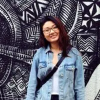 Jenny Wang stands in front of a large black and white piece of art. She is smiling, and is wearing glasses, a white shirt and light blue denim jacket.